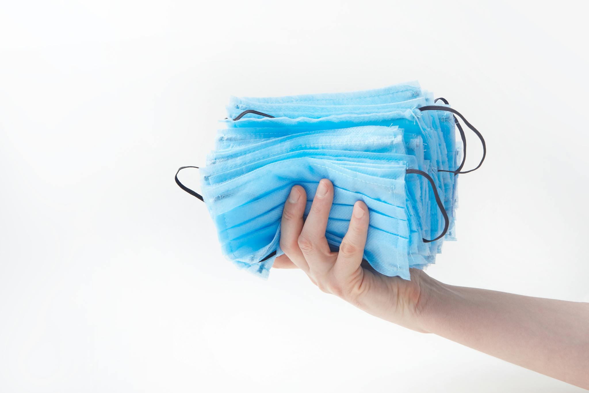 A close-up of a hand holding a stack of blue face masks against a white background.